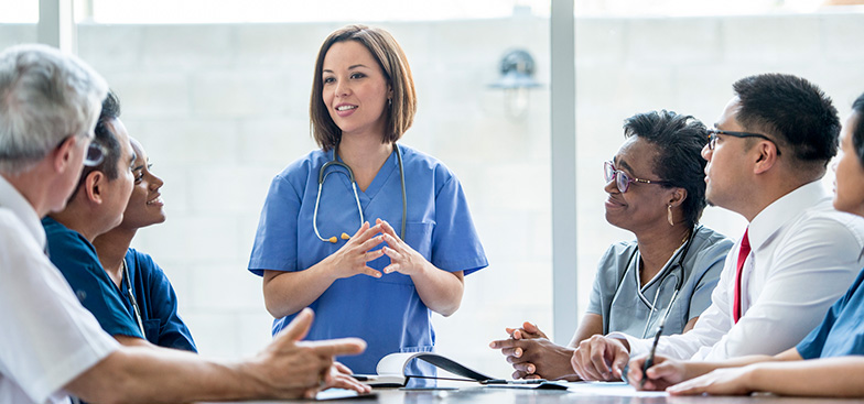 a woman wearing a stethoscope talks to people sitting around a table