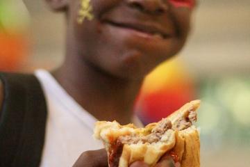 a young smiling boy with facepaint holds a cheeseburger 