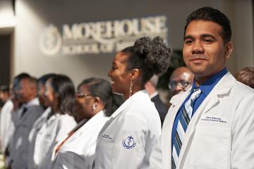 a man smiles while standing in a group of students wearing whitecoats
