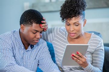 a woman feels her son's forehead while holding a tablet