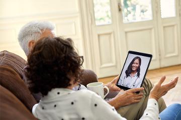 a couple sits on a couch while working with a doctor on their tablet